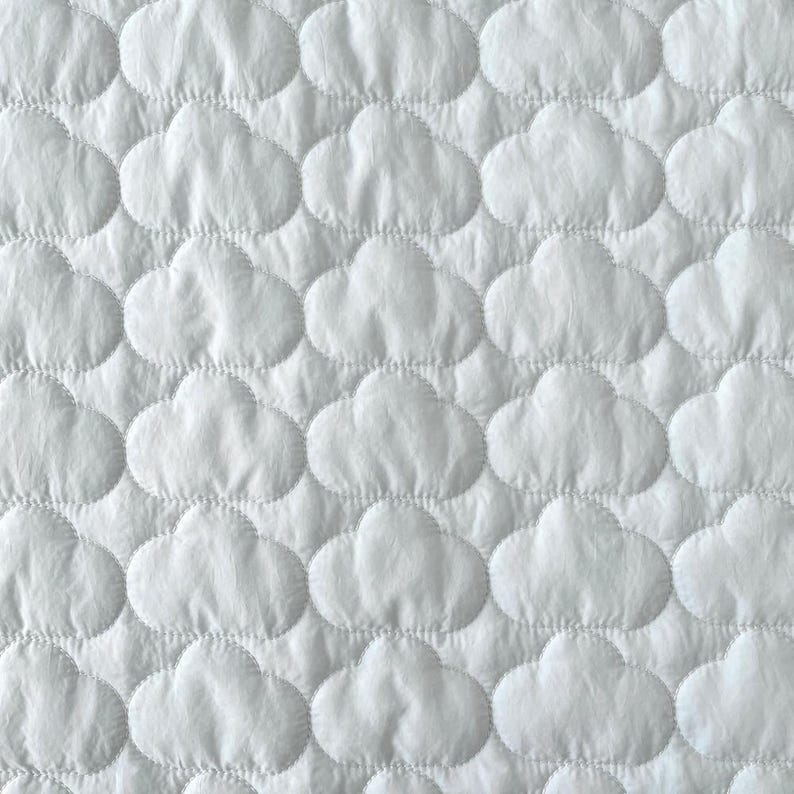 Op de afbeelding: Een witte stof met een herhalend doorgestikt patroon van bolle, afgeronde wolkvormen. Het oppervlak is getextureerd met gestikte contouren die elk individueel wolkemotief over het hele materiaal defini&euml;ren, wat zorgt voor een zachte, uniforme uitstraling.