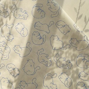 May include: White fabric with a repeating pattern of blue line-drawn cats in various poses. The cats are stylized with simple features and are scattered across the fabric. The fabric is slightly wrinkled, suggesting it is a sheet or cloth.