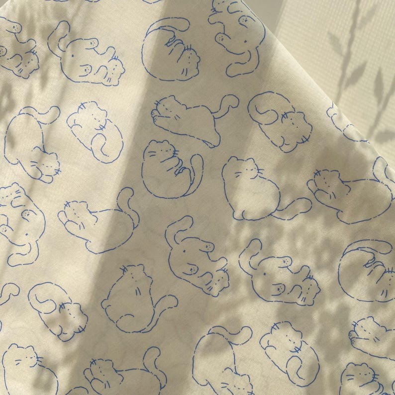 May include: White fabric with a repeating pattern of blue line-drawn cats in various poses. The cats are stylized with simple features and are scattered across the fabric. The fabric is slightly wrinkled, suggesting it is a sheet or cloth.