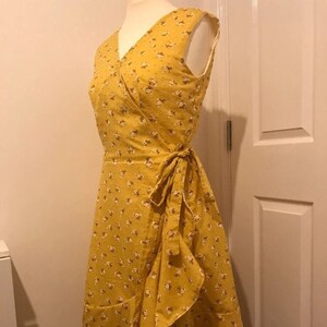 Op de afbeelding: Een gele wikkeljurk met een bloemenprint. De jurk heeft een V-hals en een strik in de taille.