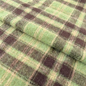 May include: A soft, fuzzy fabric featuring a classic plaid pattern in shades of light green, sage green, and dark brown. The textile has a visible nap and a textured surface, with the intersecting lines creating a repeating grid of squares.