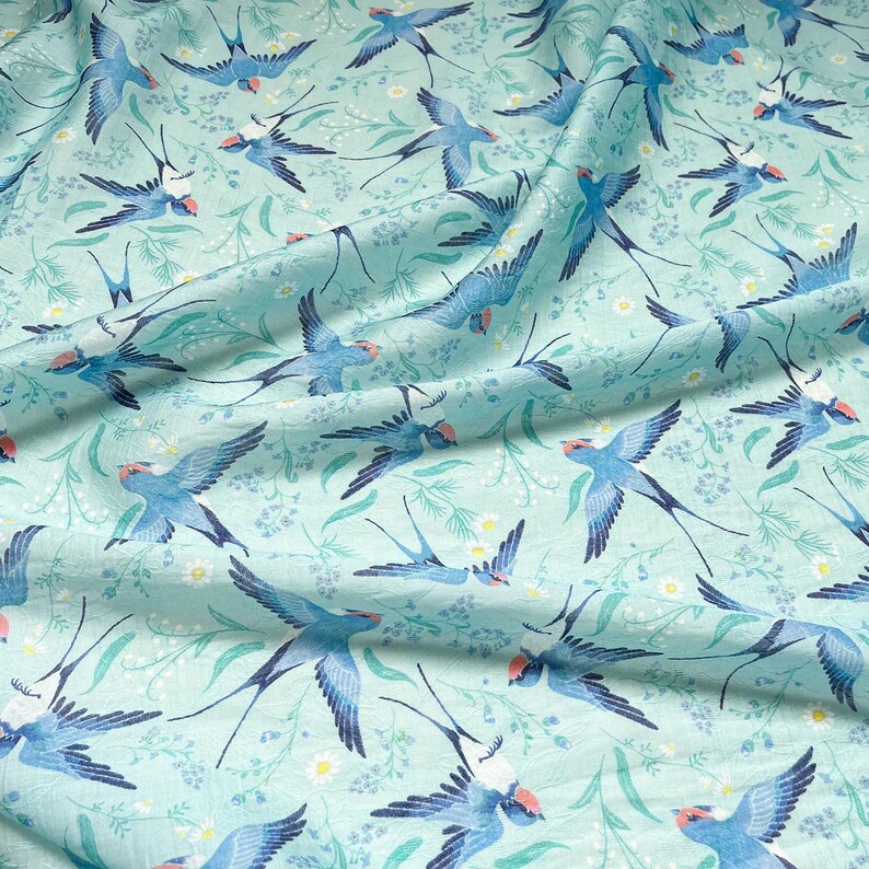 May include: A light blue fabric with a repeating pattern of blue swallows, white flowers, and green foliage. The swallows have red markings on their heads and are depicted in various flying positions. The fabric has a slightly wrinkled texture.