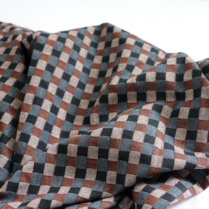 May include: Close-up of a fabric with a repeating checkered pattern. The squares alternate between brown, black, and blue-gray hues. The fabric is draped, creating soft folds and shadows, and is set against a white background.