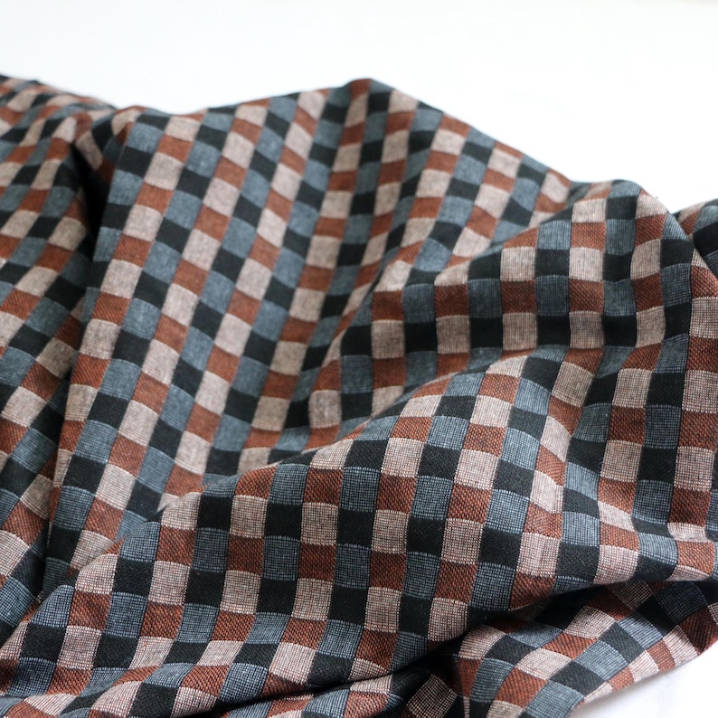 May include: Close-up of a fabric with a repeating checkered pattern. The squares alternate between brown, black, and blue-gray hues. The fabric is draped, creating soft folds and shadows, and is set against a white background.