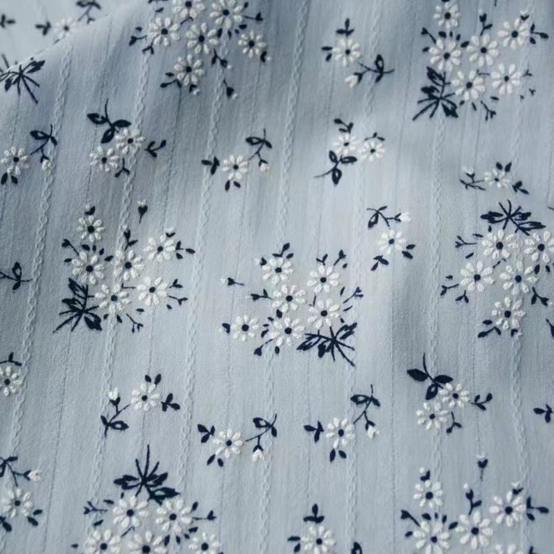 K&ouml;nnte beinhalten: Ein hellblauer Stoff mit einem wei&szlig;en Blumenmuster. Der Stoff hat eine subtile Textur und ein zartes, feminines Gef&uuml;hl.