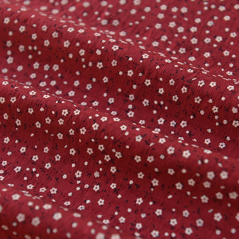 May include: Close-up of a red fabric with a delicate floral pattern. The fabric is a deep red color, and the pattern consists of small, white flowers with tiny black accents. The fabric appears to be soft and flowing, with subtle folds and wrinkles.