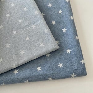 May include: Two pieces of fabric layered on top of each other. The top piece is light grey with a repeating pattern of small white stars. The bottom piece is a darker denim blue fabric, also featuring a repeating pattern of small white stars.