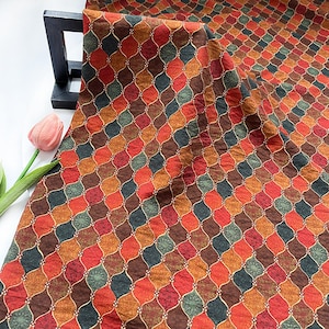 Puede incluir: Una tela estampada con un diseño de rombos repetidos en tonos de rojo, marrón, naranja y verde. La tela está drapeada sobre un soporte de madera negro, con un tulipán rosa en primer plano.