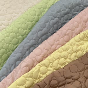 Könnte beinhalten: Eine Sammlung von gesteppten Stoffmustern mit einem sich wiederholenden Blumenmuster. Die Farben umfassen Creme, Hellgrün, Schieferblau, Zartrosa, Hellgelb und Braun. Jedes Stoffstück hat eine gesteppte Textur mit abgerundeten Blütenblättern.
