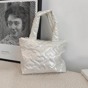 May include: A white quilted tote bag featuring a heart pattern. The bag has two handles and a rectangular shape. The bag is made of a shiny, padded material.