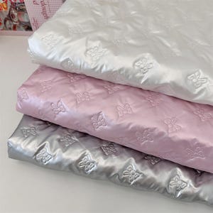 May include: Three folded layers of fabric in white, light pink, and metallic silver. Each layer features a repeating embossed pattern of small bows across the surface. The material has a smooth, slightly reflective finish with a quilted texture.