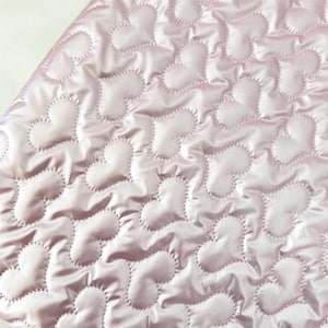 May include: Close-up of a quilted fabric featuring a repeating heart design. The material is a pale pink with a satin finish. Each heart is outlined with a contrasting stitch, creating texture and depth. The overall aesthetic is delicate and romantic, suitable for various craft projects.