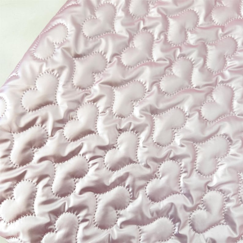 May include: Close-up of a quilted fabric featuring a repeating heart design. The material is a pale pink with a satin finish. Each heart is outlined with a contrasting stitch, creating texture and depth. The overall aesthetic is delicate and romantic, suitable for various craft projects.