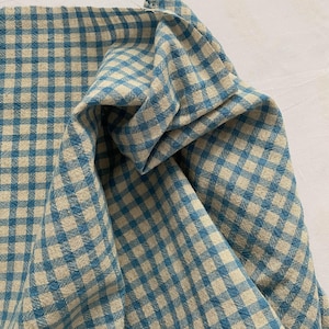 May include: Close-up of a folded fabric with a blue and beige gingham pattern. The fabric is bunched up, showing the texture and weave of the material. The pattern is a classic, small-scale check design.