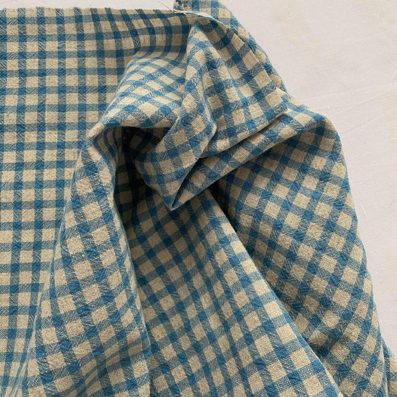 May include: Close-up of a folded fabric with a blue and beige gingham pattern. The fabric is bunched up, showing the texture and weave of the material. The pattern is a classic, small-scale check design.