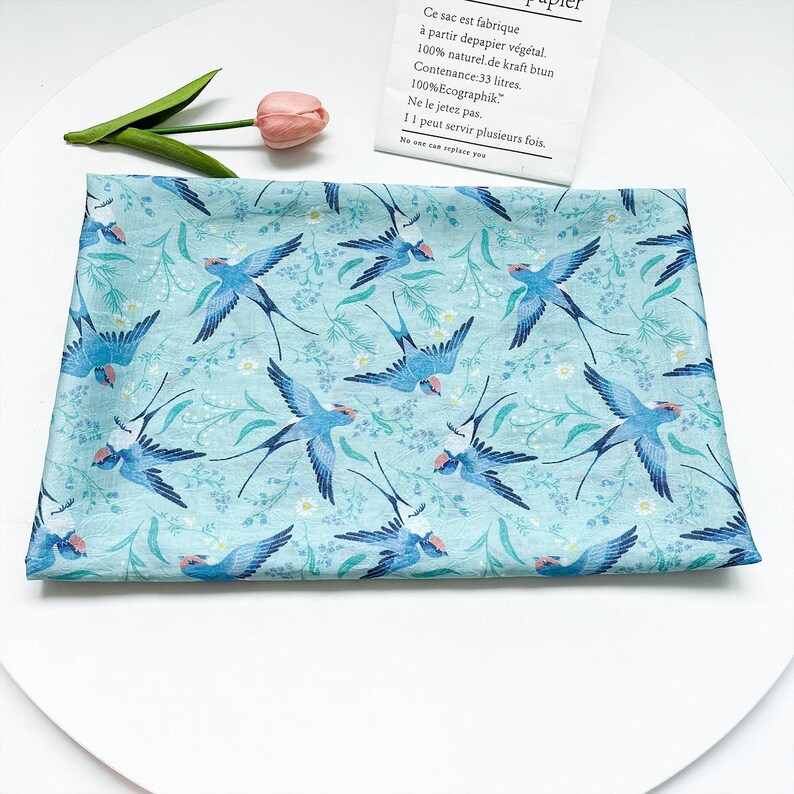 May include: A light blue fabric with a repeating pattern of blue swallows and white flowers. The swallows are in flight, with their wings spread. The fabric is likely cotton or linen, and is suitable for sewing or crafting projects.