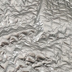 May include: A close-up of a silver quilted fabric with a diamond pattern. The fabric has a shiny, satin-like appearance, with visible folds and shadows. The diamond pattern is created by stitching, which is visible and adds to the texture and design of the fabric.