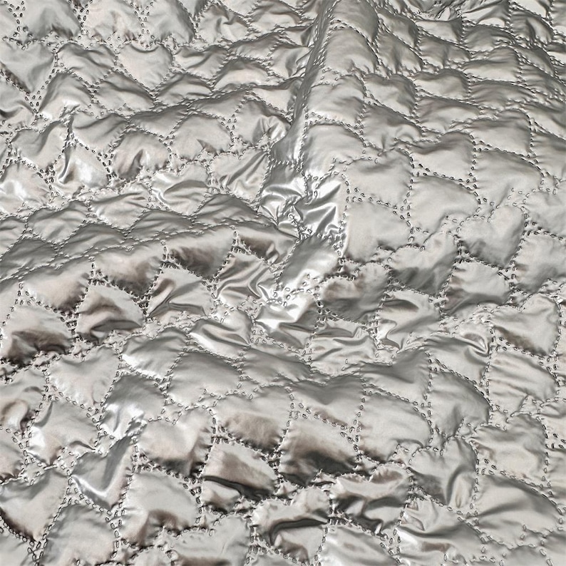 May include: A close-up of a silver quilted fabric with a diamond pattern. The fabric has a shiny, satin-like appearance, with visible folds and shadows. The diamond pattern is created by stitching, which is visible and adds to the texture and design of the fabric.