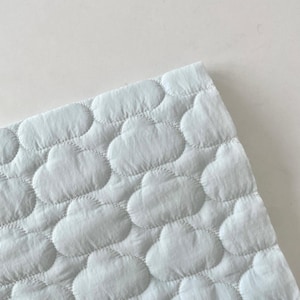 Op de afbeelding: Een lichtblauwe stof met een herhalend doorgestikt patroon van bolle, afgeronde wolkvormen. Het materiaal heeft een zachte, getextureerde uitstraling met zichtbare stiksels die elk individueel wolk-motief over het hele oppervlak omlijnen.