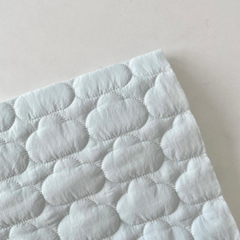 Op de afbeelding: Een lichtblauwe stof met een herhalend doorgestikt patroon van bolle, afgeronde wolkvormen. Het materiaal heeft een zachte, getextureerde uitstraling met zichtbare stiksels die elk individueel wolk-motief over het hele oppervlak omlijnen.