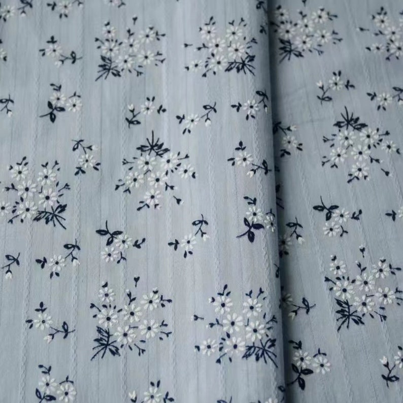 K&ouml;nnte beinhalten: Ein hellblauer Stoff mit einem wei&szlig;en Blumenmuster. Der Stoff hat eine subtile gestreifte Textur.