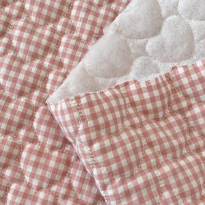 May include: A quilted fabric featuring a repeating heart-shaped pattern with a pink and white gingham check design. The material has a soft, textured surface with visible stitching outlining each heart shape, revealing a white underside.