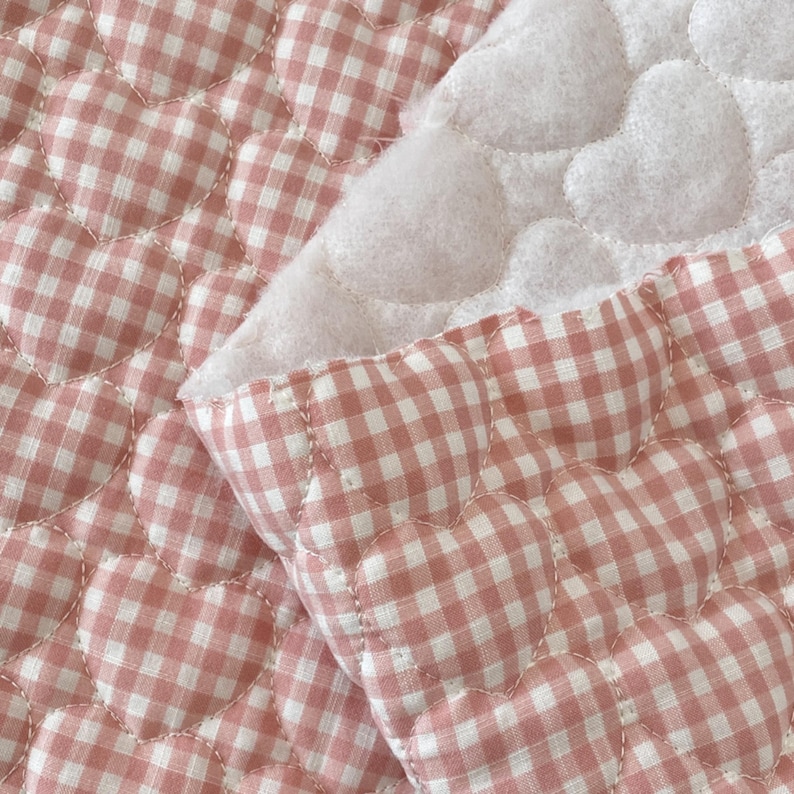May include: A quilted fabric featuring a repeating heart-shaped pattern with a pink and white gingham check design. The material has a soft, textured surface with visible stitching outlining each heart shape, revealing a white underside.