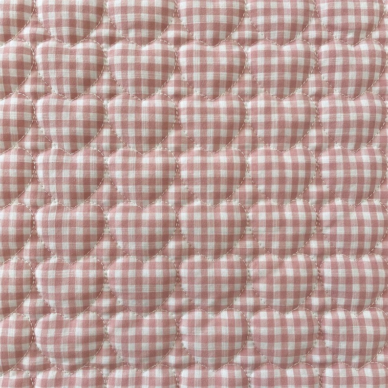 May include: A quilted fabric featuring a repeating pattern of raised, heart-shaped sections. The textile is covered in a pink and white gingham check print, with visible stitching outlining each individual heart shape across the surface.