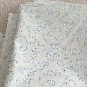 May include: White fabric with a repeating pattern of blue cat illustrations. The cats are depicted in various poses, including sitting, lying down, and curled up. The fabric is folded, showing multiple layers.