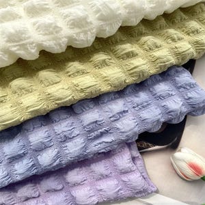 May include: A stack of four folded fabric pieces featuring a puckered, grid-like texture. The colours from top to bottom are white, light sage green, periwinkle blue, and soft lavender. Each piece displays a consistent square-patterned surface.