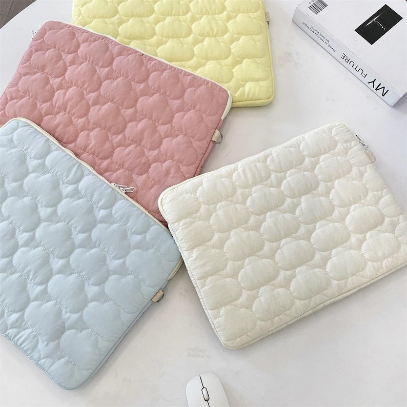 Op de afbeelding: Vier rechthoekige laptoptassen in pastelgeel, roze, lichtblauw en wit. Elke hoes heeft een gewatteerd wolkenpatroon en een ritssluiting aan de zijkant. Een wit boek met de tekst MY FUTURE en een witte computermuis zijn ook zichtbaar.