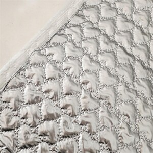 May include: A close-up of a silver quilted fabric with a repeating heart-shaped pattern. The stitching is visible, outlining each heart. The fabric has a shiny, reflective quality, and the edges are finished with a white border.