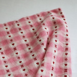 May include: Pink and white checkered fabric with a repeating pattern of red hearts and small floral designs. The fabric is folded, showing the texture and pattern. Ideal for sewing projects or crafting.
