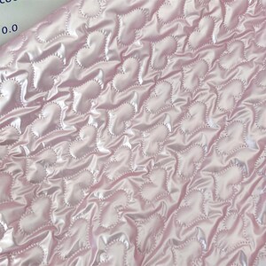 May include: Close-up of a pink satin fabric featuring a quilted heart pattern. The hearts are outlined with a dotted stitch, creating a textured effect. The fabric has a shiny, reflective quality, and the hearts are evenly spaced across the surface.