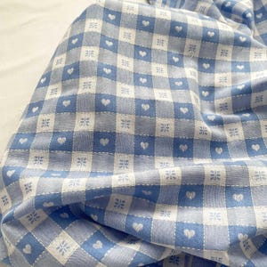 May include: A woven fabric featuring a blue and white checkered pattern. The design includes small white hearts centred within the blue squares and small white floral-like geometric motifs centred within the white squares of the grid.