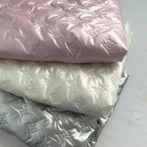 May include: Three layers of quilted fabric in pink, white, and grey. Each fabric has a glossy sheen and a repeating embossed butterfly design. The fabrics are folded and stacked, creating a layered effect.