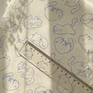 May include: White fabric with a repeating pattern of blue cat illustrations. A clear ruler with centimeter markings is placed diagonally across the fabric, showing measurements up to 15 cm.