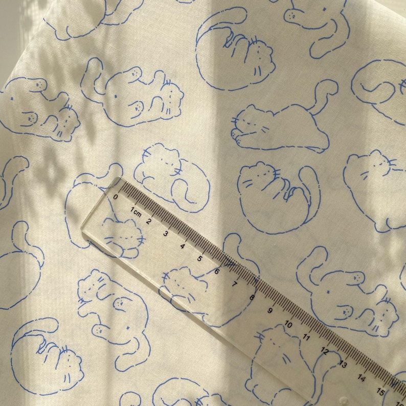 May include: White fabric with a repeating pattern of blue cat illustrations. A clear ruler with centimeter markings is placed diagonally across the fabric, showing measurements up to 15 cm.