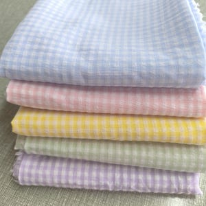 Könnte beinhalten: Ein Stapel von sechs pastellfarbenen Stoffen mit Vichy-Muster. Die Farben umfassen Hellblau, Rosa, Gelb, Grün und Lila. Der Stoff wirkt weich und leicht, mit einer leicht zerknitterten Textur.