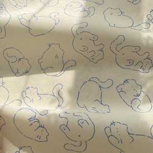 May include: Off-white fabric with a repeating pattern of blue cat illustrations. The cats are depicted in various playful poses, with simple line drawings creating a whimsical design. The fabric has a soft, slightly textured appearance.