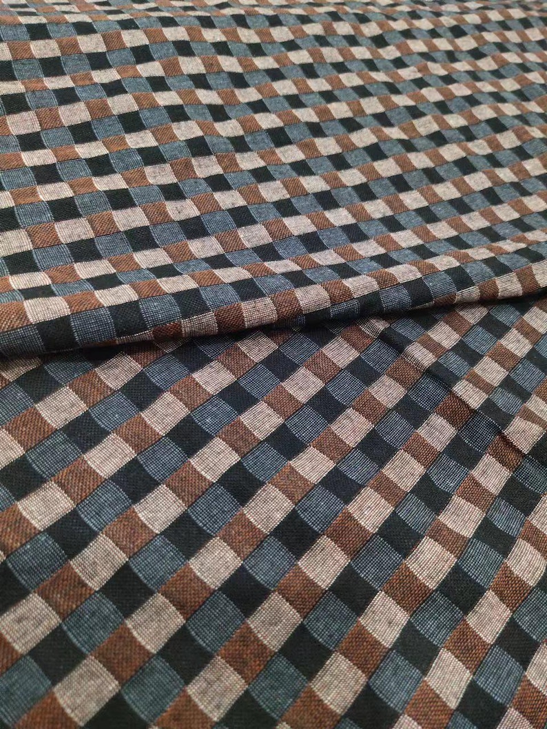 May include: Close-up of a fabric with a checkered pattern. The pattern features squares in shades of brown, black, blue, and tan. The fabric is folded over, showing the texture and weave of the material. The overall appearance is of a woven textile.