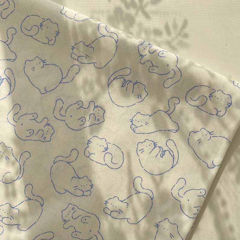 May include: White fabric with a repeating pattern of blue cat illustrations in various poses. The cats are drawn with simple outlines, creating a playful and whimsical design. The fabric appears to be soft and lightweight.
