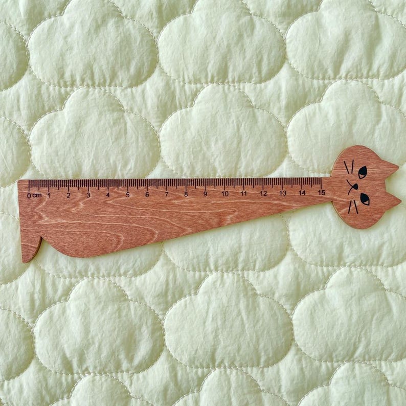 Op de afbeelding: Een houten liniaal in de vorm van een kat, met een natuurlijke houtnerf en een zwart bedrukt kattengezicht aan de rechterkant. De liniaal toont duidelijke meetmarkeringen van 0 tot 15 centimeter langs de bovenrand voor nauwkeurige metingen.