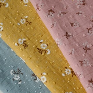 Op de afbeelding: Drie stofstalen met een wit bloemenpatroon op een textuur achtergrond. De stalen zijn in de kleuren blauw, geel en roze.