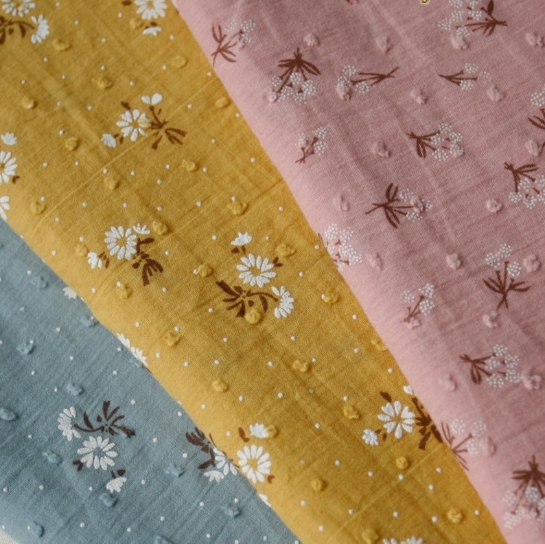 Op de afbeelding: Drie stofstalen met een wit bloemenpatroon op een textuur achtergrond. De stalen zijn in de kleuren blauw, geel en roze.