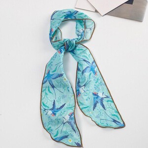 May include: A light blue scarf with a bird and floral pattern. The scarf features a repeating design of blue birds and green leaves on a light blue background. The edges are trimmed with a brown border.