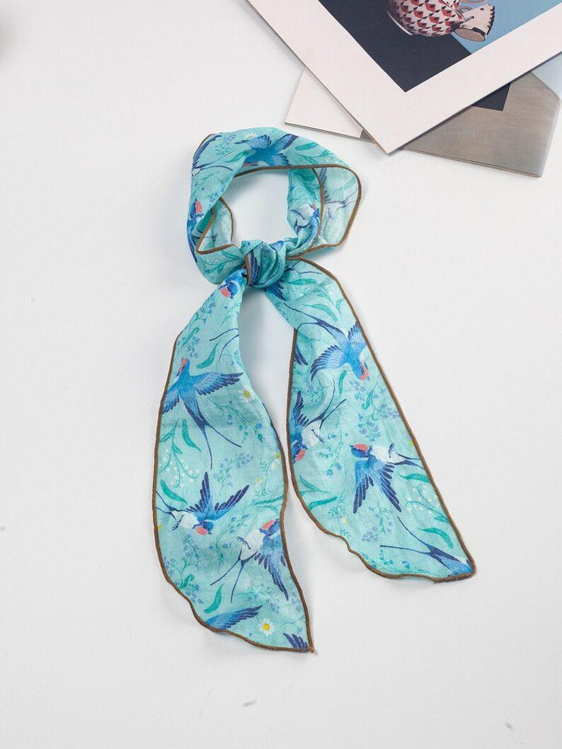 May include: A light blue scarf with a bird and floral pattern. The scarf features a repeating design of blue birds and green leaves on a light blue background. The edges are trimmed with a brown border.