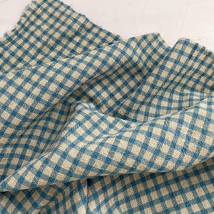 May include: A close-up of a blue and beige gingham fabric. The fabric is folded and draped, showcasing the pattern's texture and the interplay of colors. The fabric has a soft, slightly wrinkled appearance.