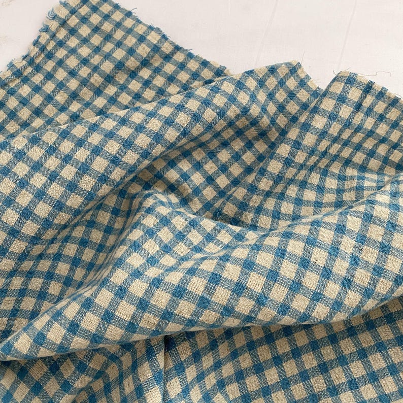 May include: A close-up of a blue and beige gingham fabric. The fabric is folded and draped, showcasing the pattern's texture and the interplay of colors. The fabric has a soft, slightly wrinkled appearance.