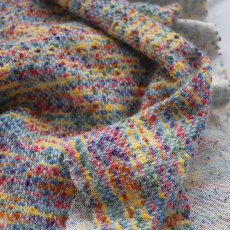 May include: A textured knit fabric featuring a multicolored speckled pattern with vibrant hues of yellow, blue, pink, orange, and purple. The material has a soft, fuzzy appearance with a dense weave, showing a folded drape that highlights the depth.